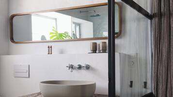 Une salle de bain dotée d’un grand lavabo ovale, d’un miroir avec un cadre en bois et d’un espace douche visible à travers une porte en verre.