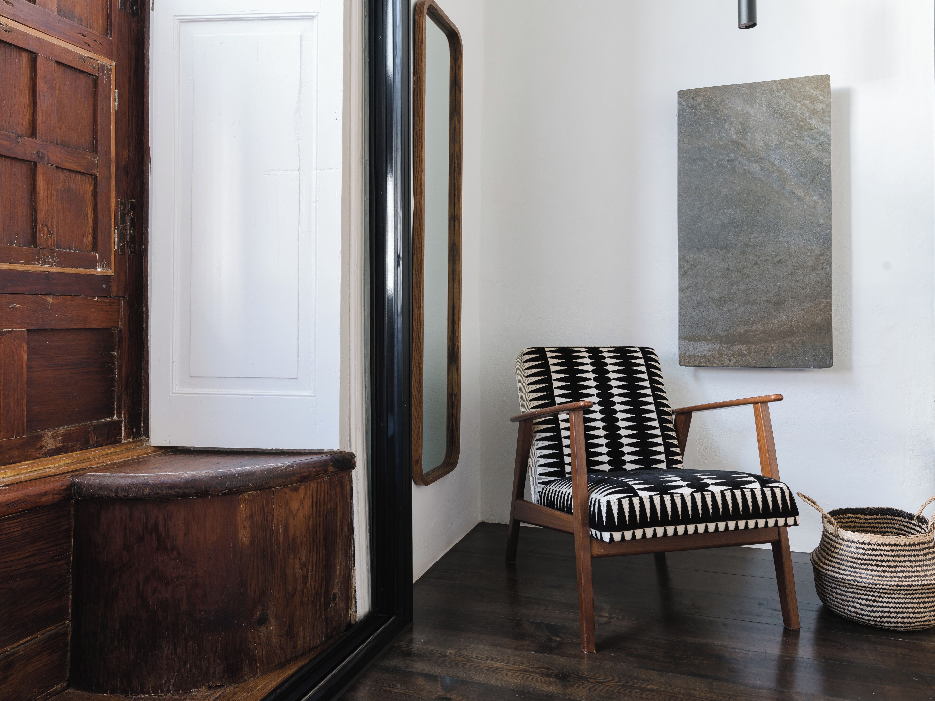 Une porte en bois, un mur blanc, une chaise à motifs noir et blanc, un panier tressé et un tableau abstrait gris.