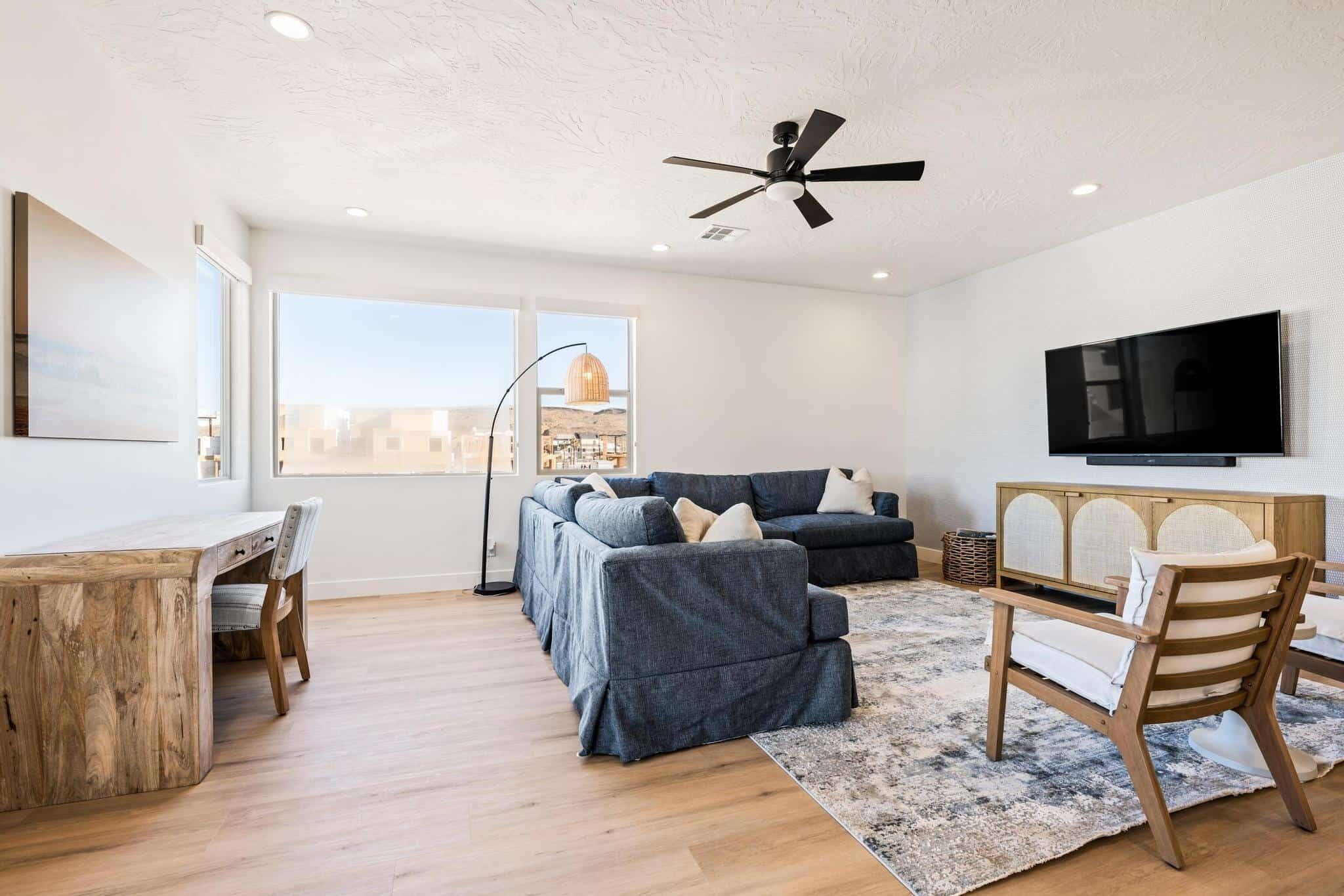 A modern living room with a sectional sofa, a wooden desk, a flat-screen TV, and a ceiling fan.