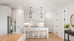 A modern kitchen with white cabinetry, a central island with stools, and a dining area with a wooden table and white chairs. - Somewhere on a Beach: Desert Color Lagoon + Beaches (St. George)
