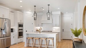 A modern kitchen with white cabinetry, stainless steel appliances, a central island with stools, and pendant lighting. - Somewhere on a Beach: Desert Color Lagoon + Beaches (St. George)