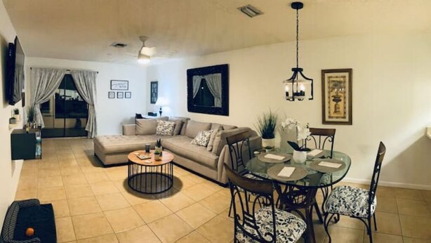 A living room with a sofa, coffee table, and dining table with chairs. There is a TV mounted on the wall and a window with curtains. - Lovely 1 bedroom unit with pool (Sebring)