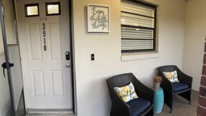 A front door with a welcome mat, two wicker chairs with cushions, a window with blinds, and a wall-mounted sign. - Lovely 1 bedroom unit with pool (Sebring)