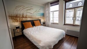 Una cama bien hecha con una colcha blanca y dos almohadas anaranjadas, un mural en la pared, una lámpara de pie y una ventana con vistas a edificios.