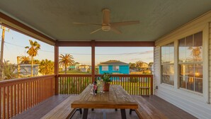 Een houten terras met een tafel en banken, een plafondventilator en een uitzicht op een blauw huis op de achtergrond.