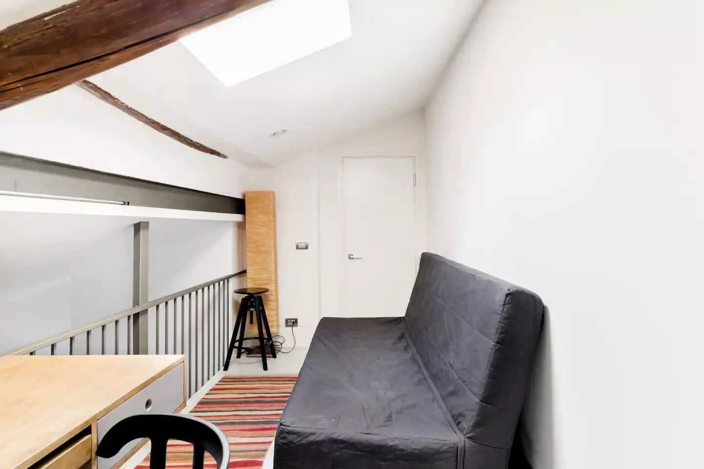 A small, compact room with a bed, a desk, and a chair. There is a window providing natural light, and a door leading to another room.