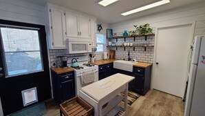 Une cuisine compacte avec des meubles blancs, un four à micro-ondes, une cuisinière et un évier. On y trouve des étagères remplies de vaisselle et une plante en pot.