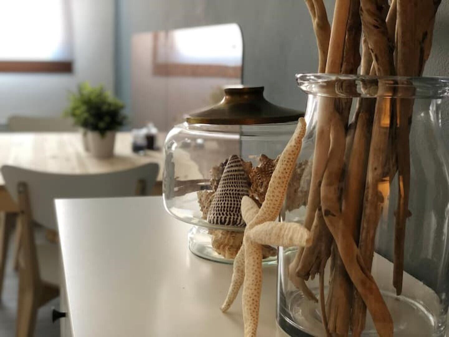 A glass jar with a lid containing a small decorative item and a plant, placed on a white table in a room with a window and a plant in the background.