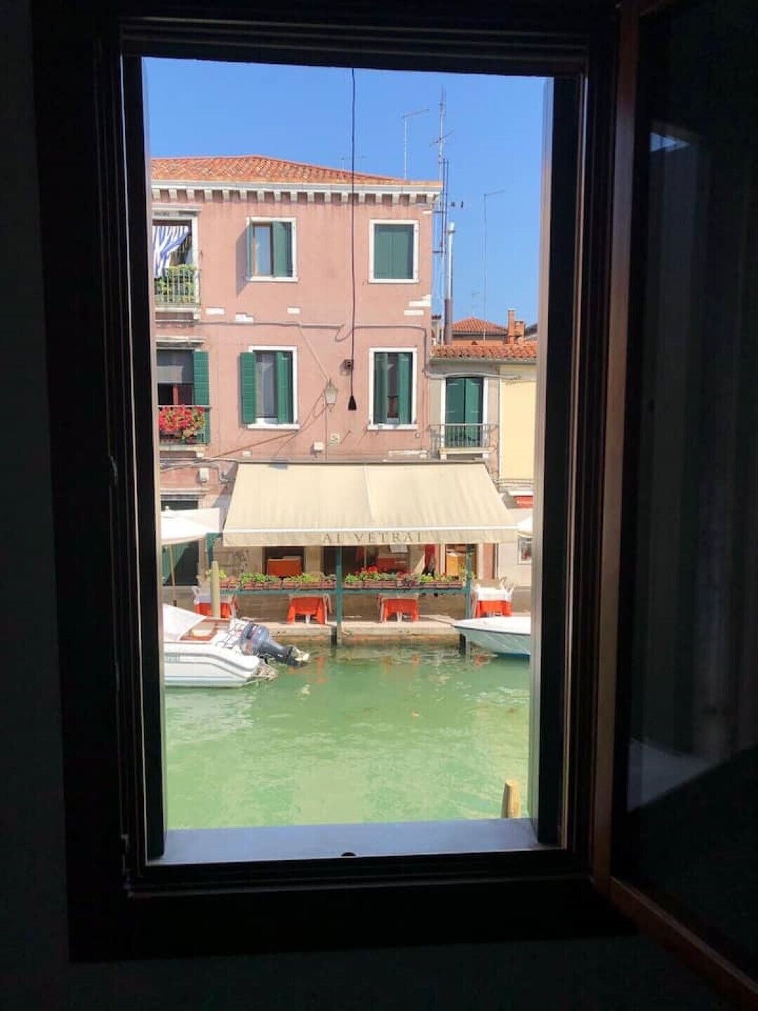 A view through a window of a building with a balcony, a boat docked in front, and a building with a sign that reads "AI VETRAI".