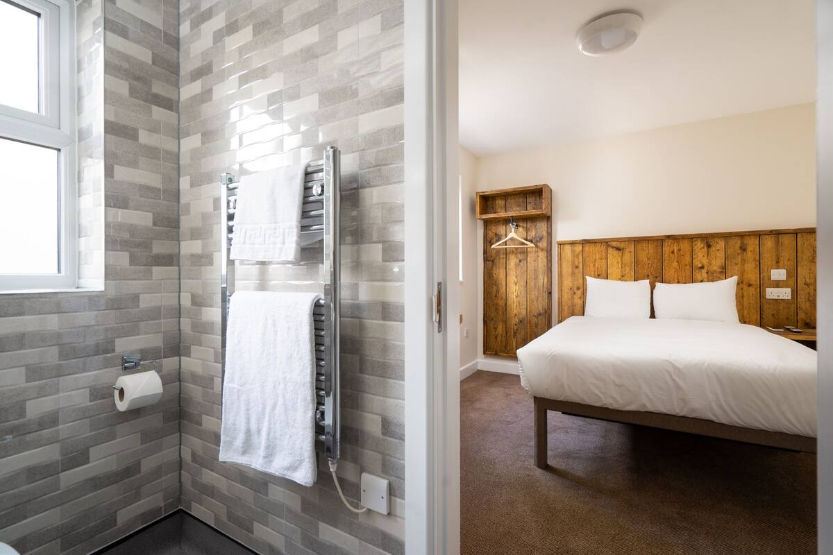 A bathroom with a glass shower enclosure, a towel rack, and a toilet paper holder. The room features a wooden headboard and a bed with white bedding.