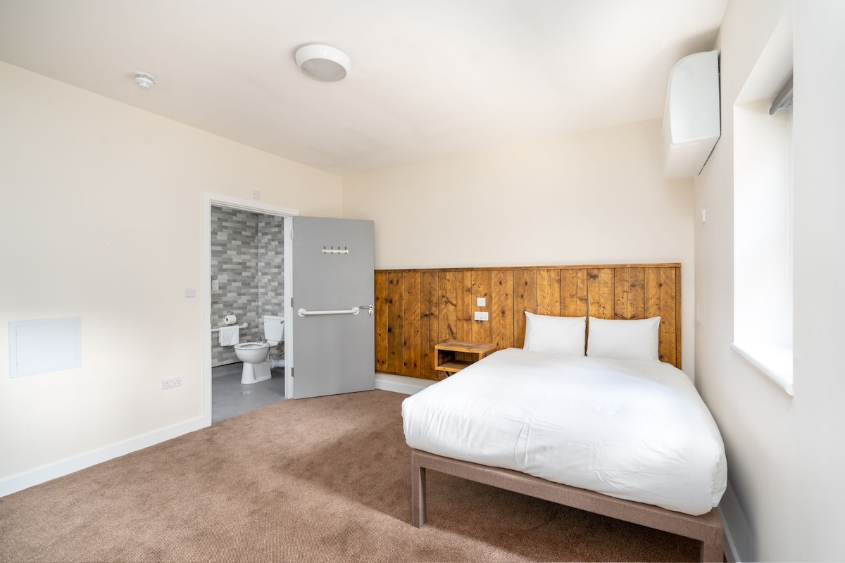 A bedroom with a wooden headboard, a bed, a small bedside table, and a bathroom visible through an open door.