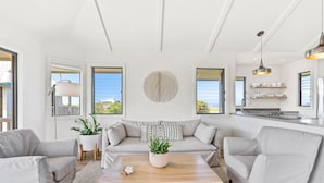 Een moderne woonkamer met een bank, twee fauteuils, een salontafel en een potplant. Er zijn grote ramen met zonwering, een keuken met een spoelbak en planken, en een plafond met zichtbare balken.