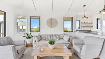 Un salon moderne comprenant un canapé, deux fauteuils, une table basse et une plante en pot. De grandes fenêtres sont équipées de stores, et on y trouve un coin cuisine avec un évier et des étagères, ainsi qu'un plafond avec des poutres apparentes.