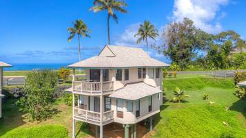 Une maison à deux étages dotée d’un balcon, entourée d’une végétation luxuriante et de palmiers, offrant une vue dégagée sur un ciel bleu azur et l’océan.
