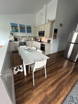 A modern kitchen with white cabinetry, a stainless steel refrigerator, and a dining area with a white table and chairs. - Lakeside three bedroom boathouse. (Wasilla)
