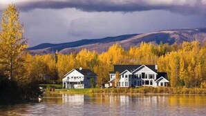 A two-story house with a large front porch, situated by a lake with autumn-colored trees in the background. - Lakeside three bedroom boathouse. (Wasilla)
