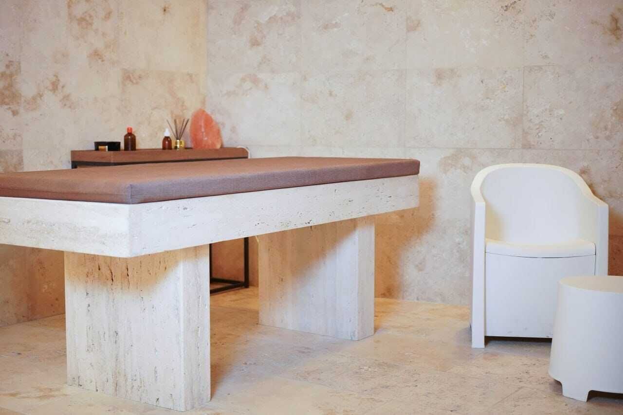 A spa room with a massage table, a white chair, and a small table with candles and a diffuser.