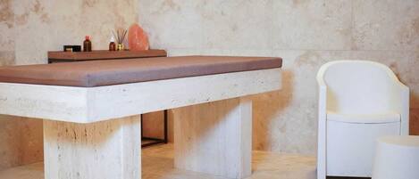 A spa room with a massage table, a white chair, and a small table with candles and a diffuser.