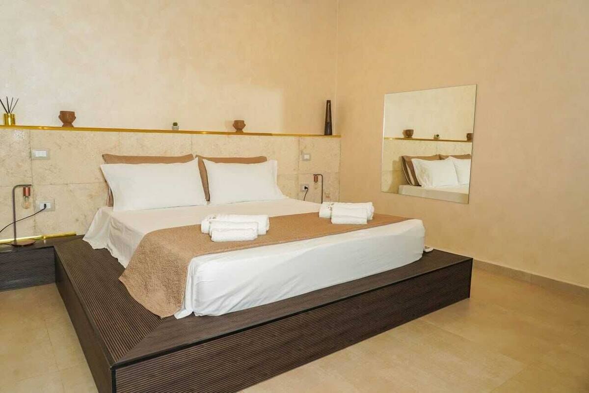 A modern hotel room with a large bed, a bedside table, a mirror, and a shelf with decorative items.