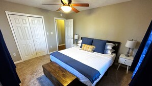 Un dormitorio con cama, mesitas de noche, un banco, ventilador de techo y una puerta.