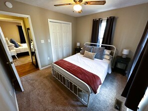 A bedroom with a bed, a nightstand, a ceiling fan, a door, and a carpeted floor.