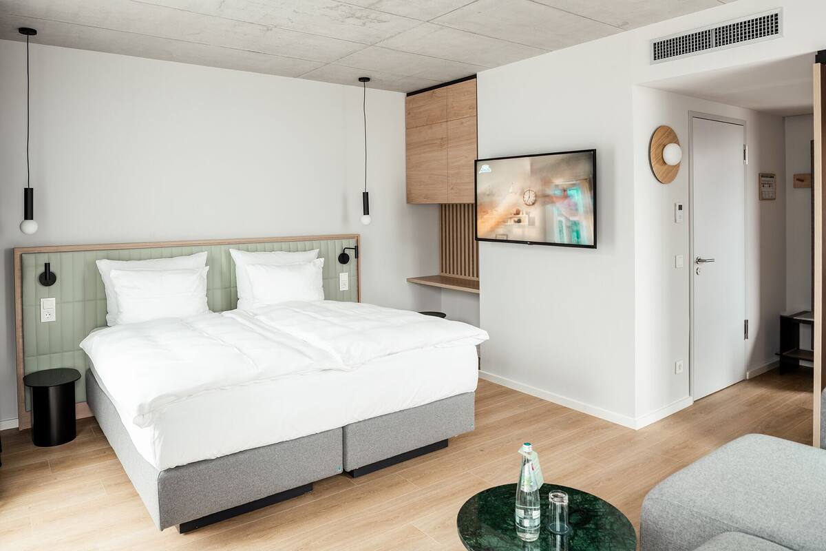 A modern bedroom with a large bed, a bedside table, a wall-mounted TV, and a door leading to another room.