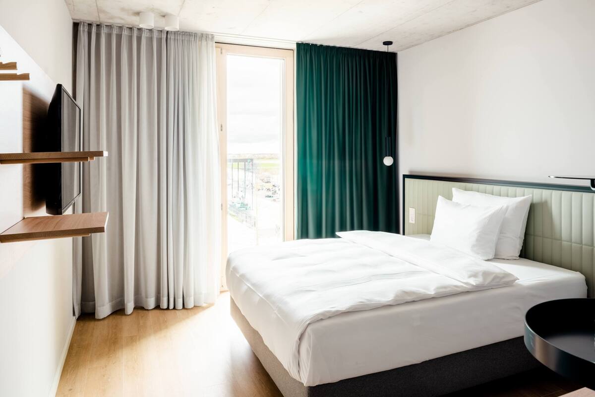 A hotel room with a bed, a TV on a wooden shelf, a balcony with curtains, and a view of the outside.