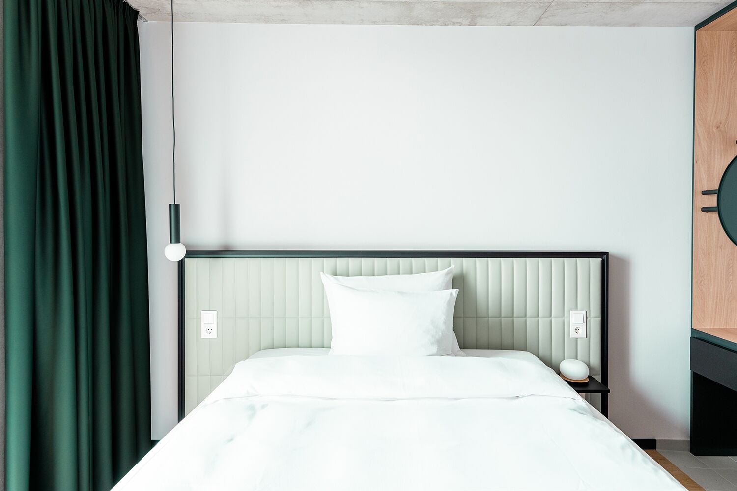 Een opgemaakt bed met witte lakens, een groen hoofdeinde en een nachtkastje met een lamp.