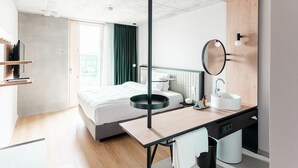 A modern hotel room with a bed, a desk with a mirror, a sink, and a view of the city through the window. - Coreum Hotel (Stockstadt am Rhein)