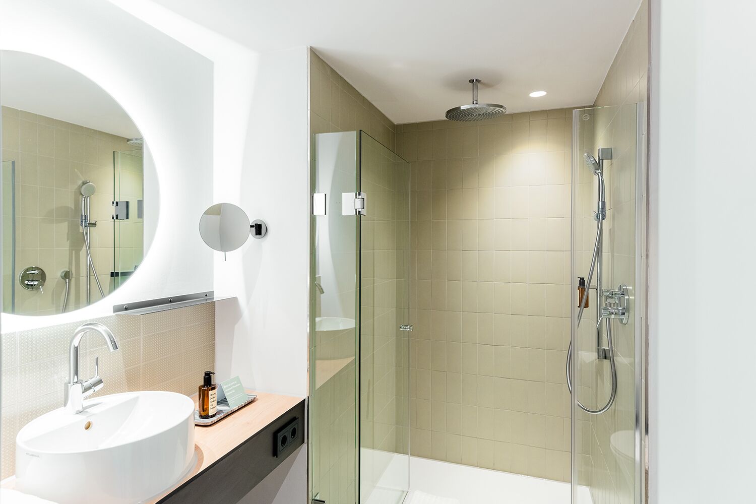 Un baño moderno con un espejo redondo, un lavabo blanco, una cabina de ducha de vidrio y un cabezal de ducha de mano.