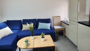 Una sala de estar moderna con un sofá azul, una mesa de madera con plantas, una silla de madera y una cocina con armarios.