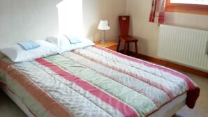 Een slaapkamer met een bed, een stoel, een bureau, een lamp, een raam met gordijnen en een radiator.