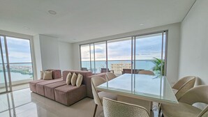 A spacious living area with a glass dining table, cushioned seating, and large windows offering a view of the sea and buildings. - LivingCartagena  (Cartagena)