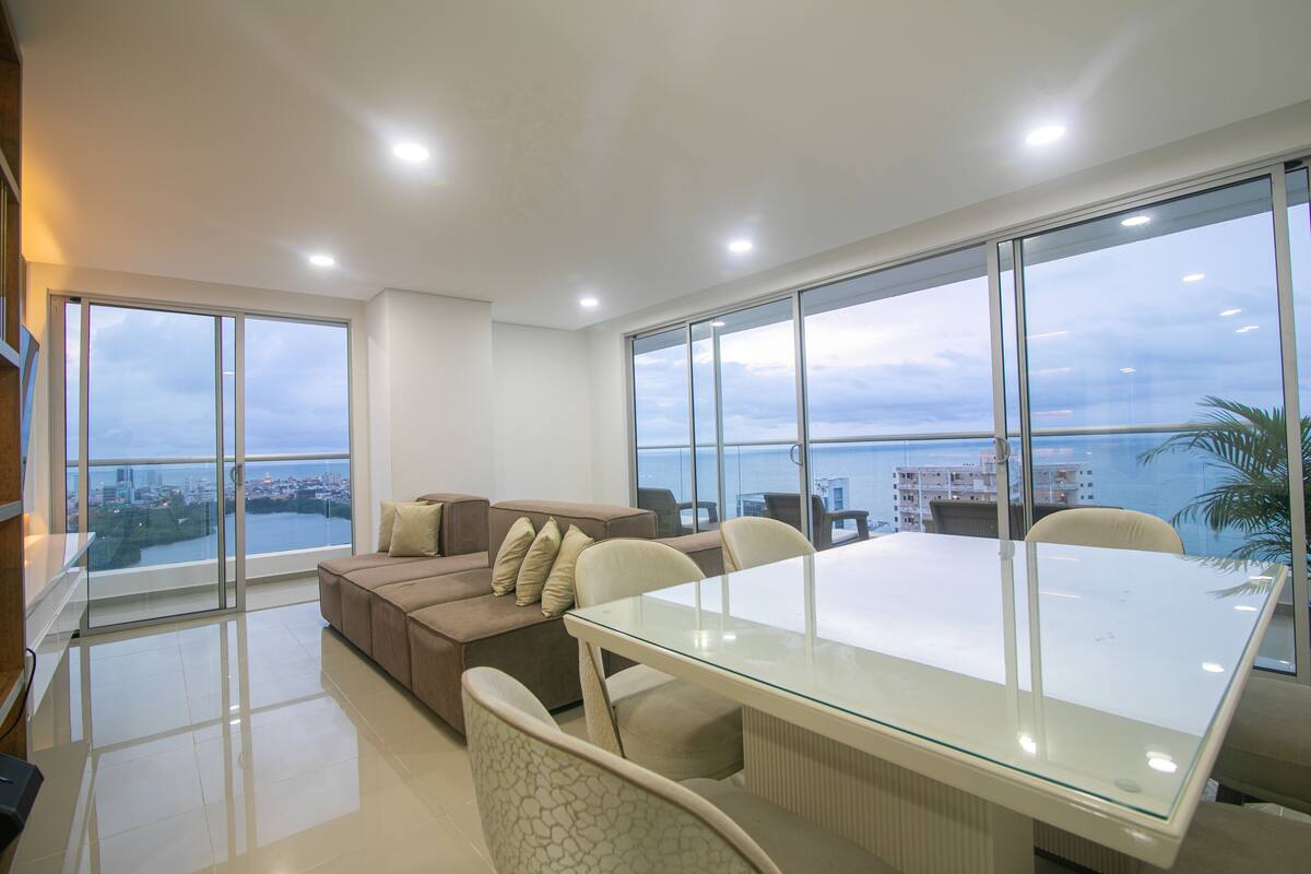 A modern living room with a glass table and sofa, large windows with a city view, and a balcony with a view of the sea.