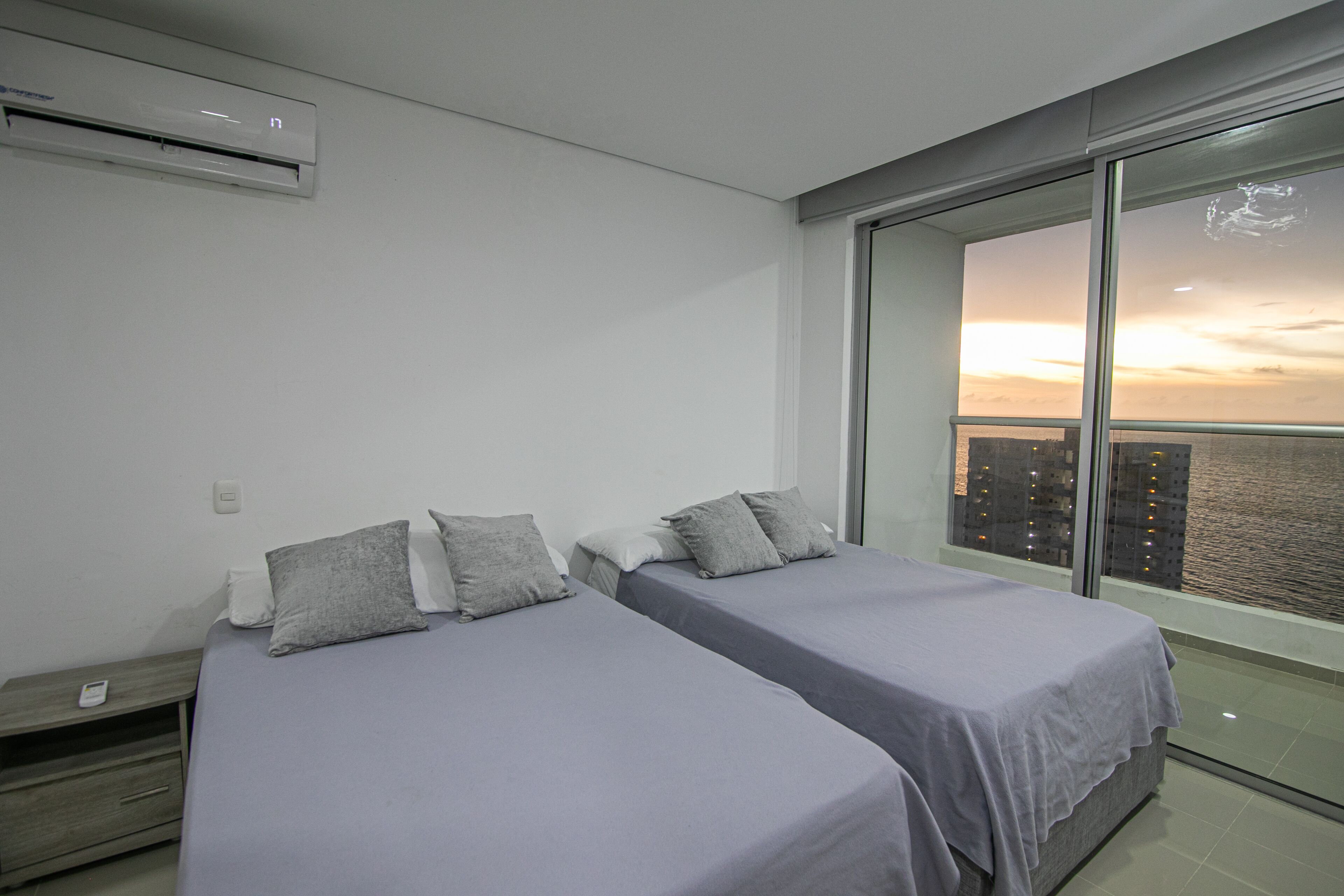 A modern hotel room with two beds, a large window with a city view, and an air conditioning unit on the wall.
