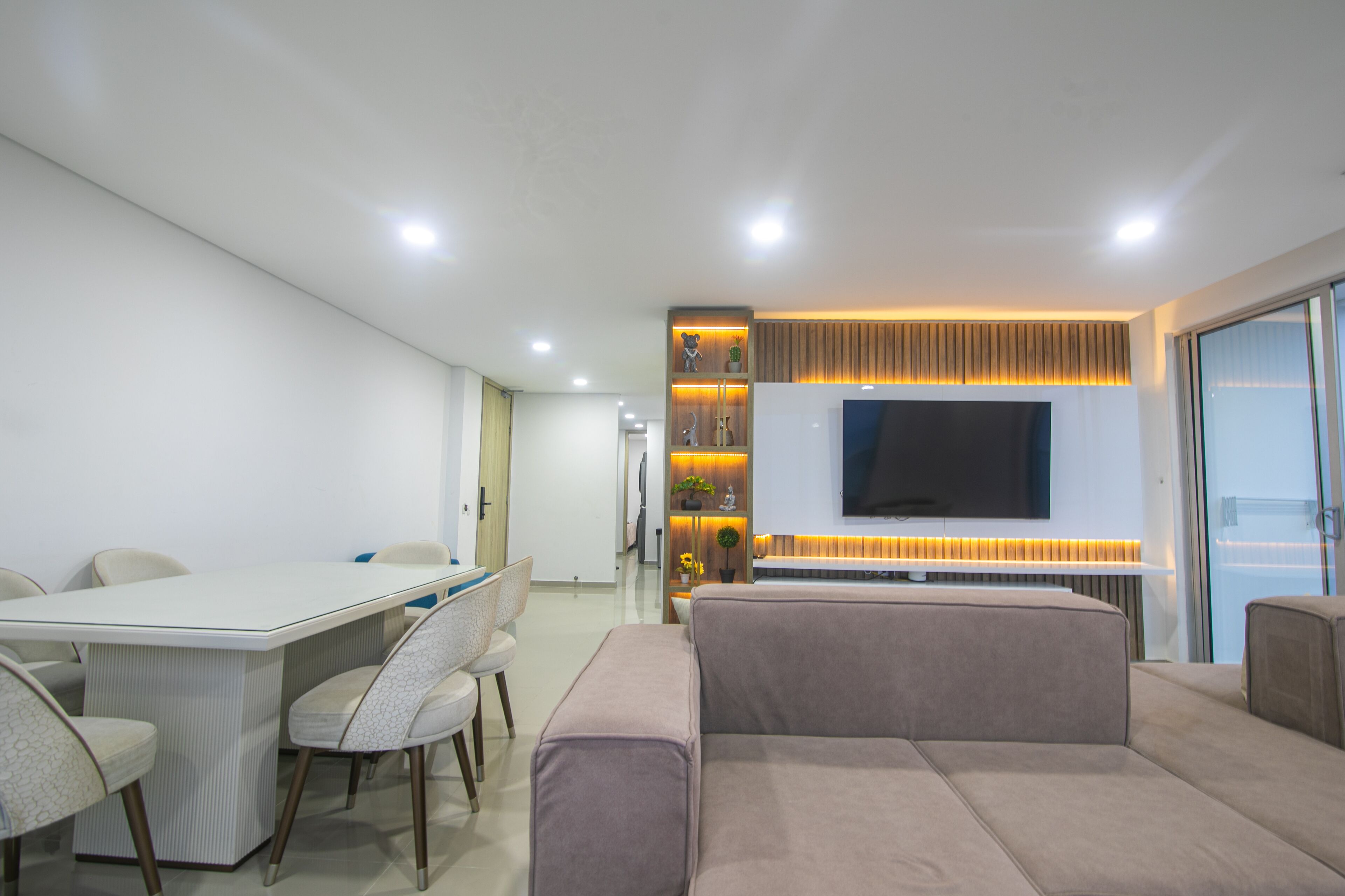 A modern living room with a flat-screen TV, a sofa, a dining table with chairs, and a built-in entertainment unit with shelves.
