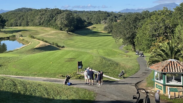 Ein Golfplatz mit Teich, Clubhaus und einer Straße mit Personen und Golfcarts.