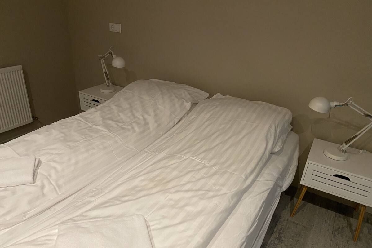 A single-bed room with white bedding, two bedside tables with lamps, and a radiator on the left.