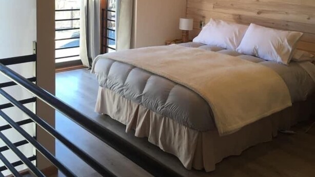 Un dormitori amb sostre de fusta, un llit amb roba de llit blanca, una nitzeta amb una làmpada i una finestra amb cortines.