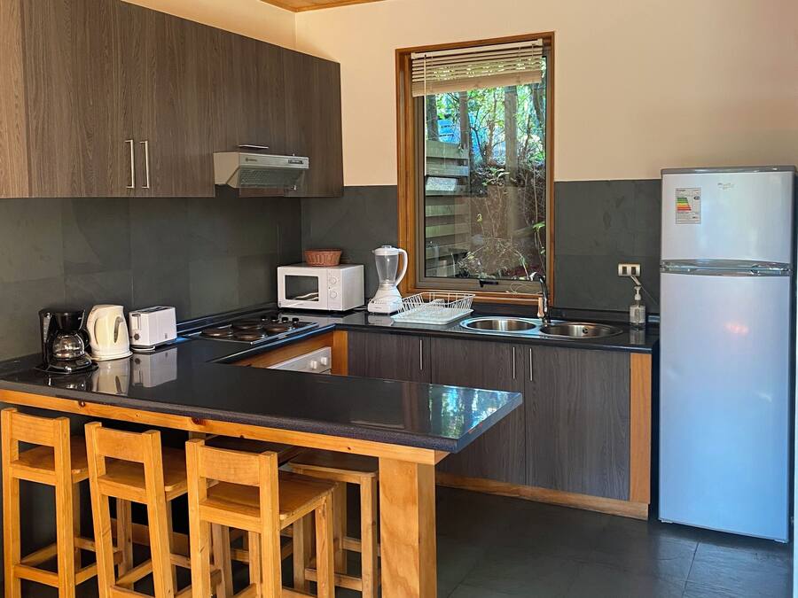 A modern kitchen with a central island, wooden bar stools, dark cabinetry, a stainless steel sink, and a built-in microwave.