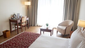 A hotel room with a bed, a desk, a chair, a small table, and a lamp.