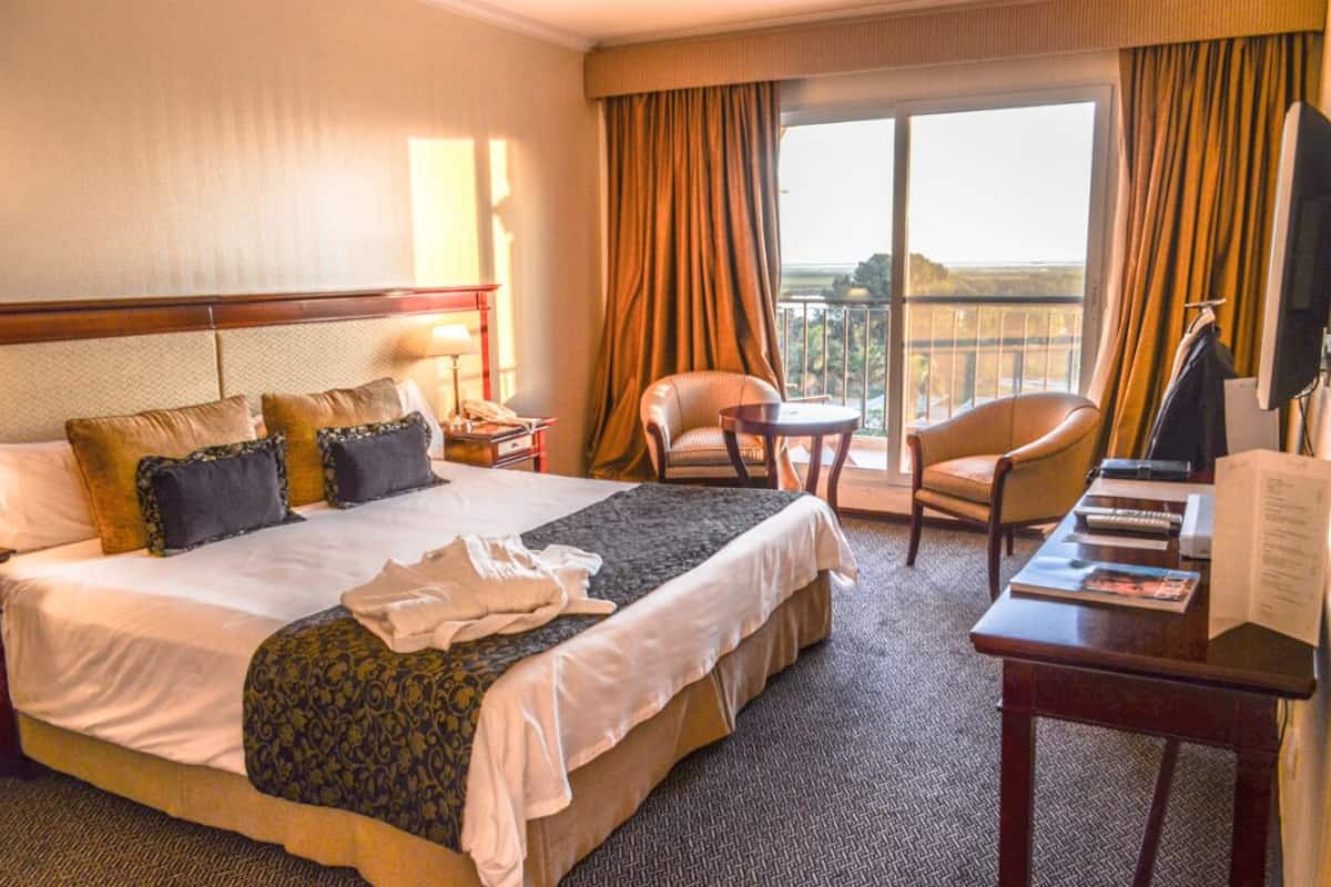 A hotel room with a large bed, a desk, a chair, and a balcony with curtains.