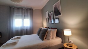En pænt redt seng med hvid dyne og flere puder, en senget lampe og et vindue med lette gardiner.