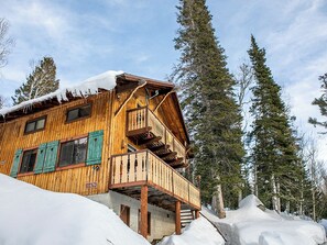 Una cabaña de madera con balcón, rodeada de nieve y altos pinos.