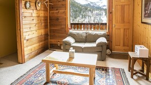 Une pièce chaleureuse aménagée dans un chalet, avec des murs en bois, une fenêtre donnant sur les montagnes, un canapé, une table et un petit tapis.