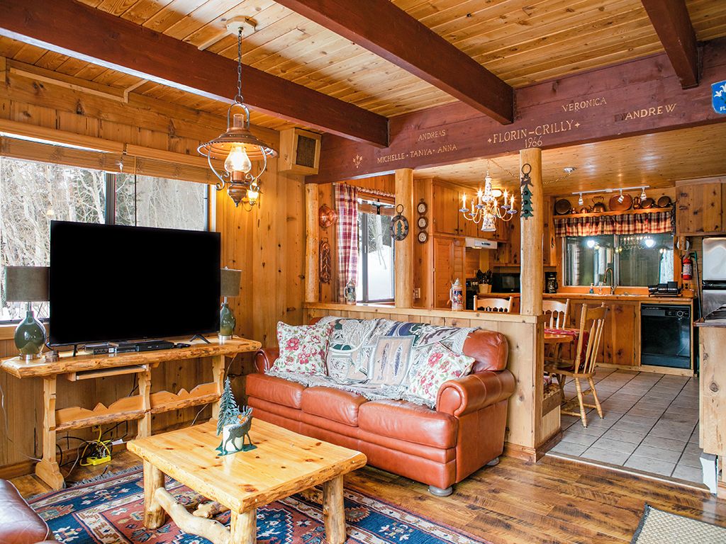 A cozy cabin interior with a wooden dining table, a leather sofa, a television, and a kitchen area.