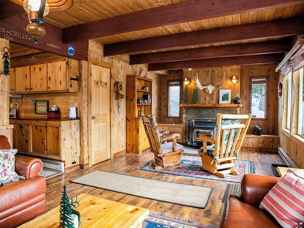 Interior rústico de una cabaña con paredes de madera, chimenea, comedor y un rincón de descanso acogedor.