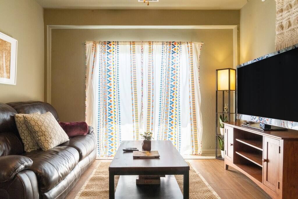 A living room with a brown leather sofa, a wooden coffee table, a TV on a wooden stand, and a patterned curtain.