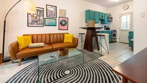 Una sala de estar moderna con un sofá de cuero marrón, una mesa de centro de vidrio y un tapete con diseño blanco y negro. Hay una zona de cocina con armarios verde azulado y un cartel de bienvenida.