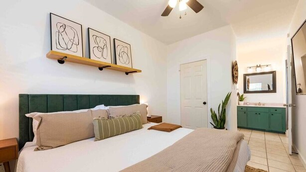 Un dormitorio con una cama grande, un cabecero verde, una mesita de noche y un velador. Se ve un baño con un lavabo y armarios.